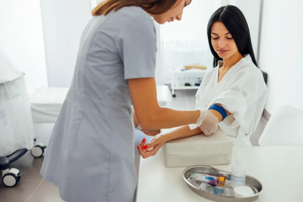 Phlebotomist preparing to draw blood from a patient using a blue tourniquet in a medical clinic, with blood collection tubes on a tray.