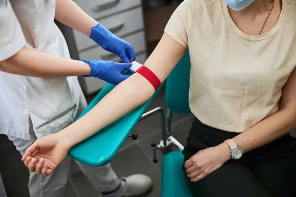 Phlebotomist applying a red tourniquet to a patient's arm before drawing blood in a clinical setting.