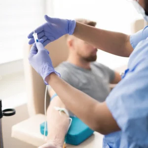 Healthcare professional in blue gloves preparing IV drip for male patient during intravenous therapy session in medical clinic.