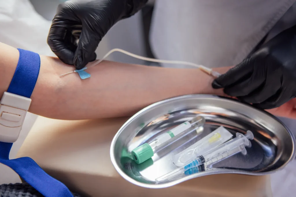 Healthcare professional wearing black gloves inserting IV catheter into patient’s arm for blood draw, with tourniquet, syringes, and blood collection tubes on medical tray in clinical setting