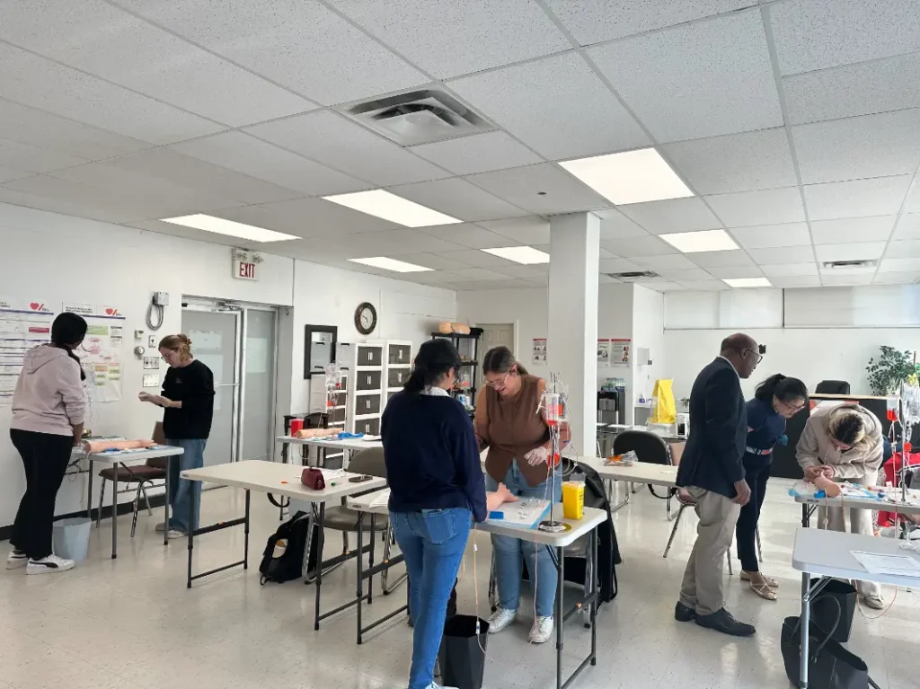 Wide classroom shot with multiple groups practicing IV/phlebotomy skills across the room.