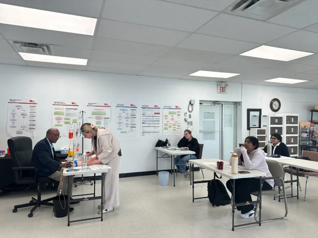 Medical training classroom with instructor demonstrating IV insertion on simulation arm while students observe at desks