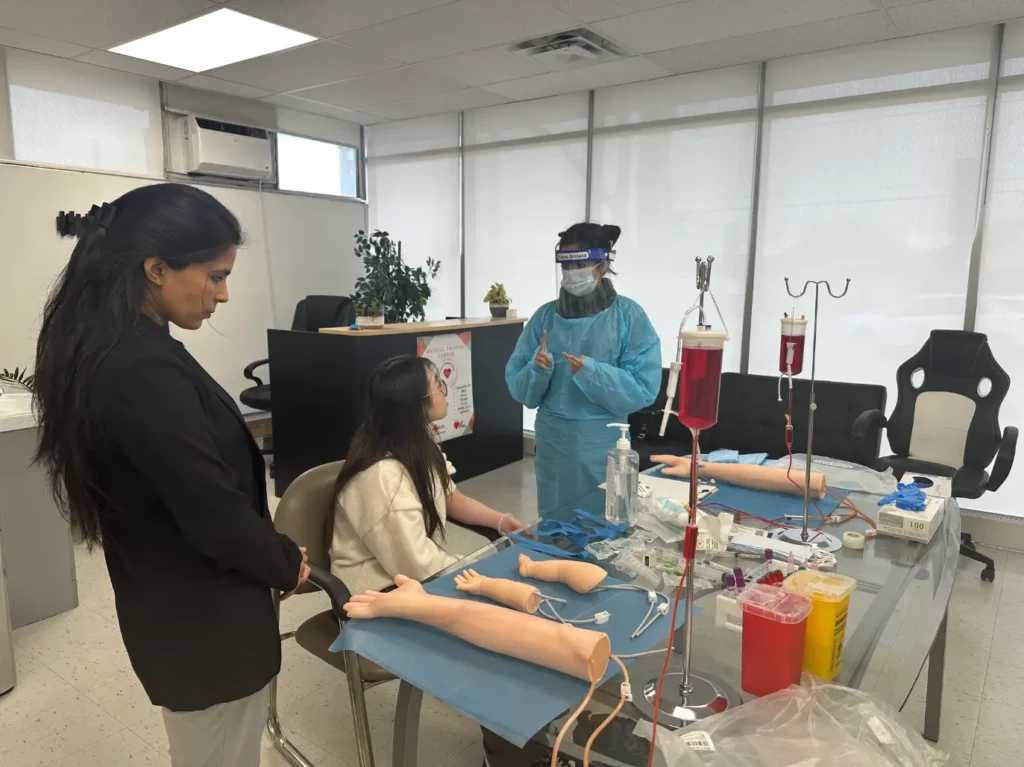 Instructor in full PPE (mask, face shield, gown) teaching IV setup with simulation arms on a table.