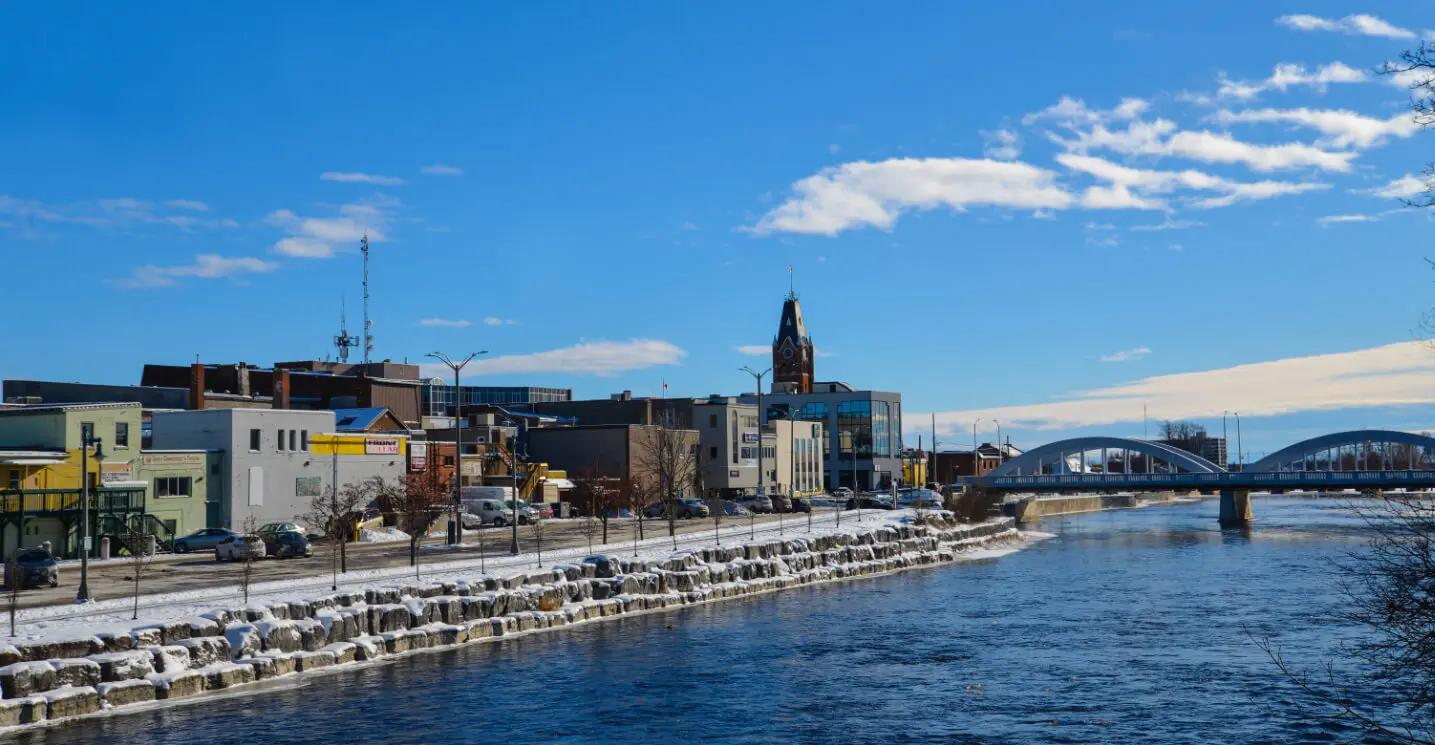 Belleville Ontario waterfront cityscape with Moira River, arched bridge, snow-covered shoreline, and downtown buildings under clear blue winter sky