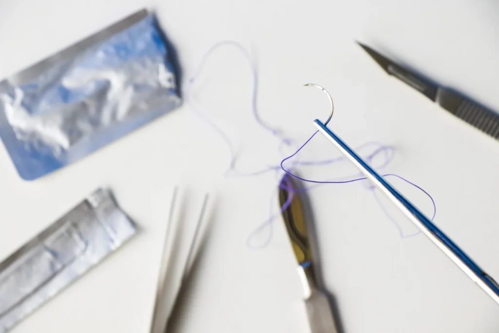 Close-up of surgical needle holder with curved suture needle and thread, surrounded by sterile medical instruments on white background