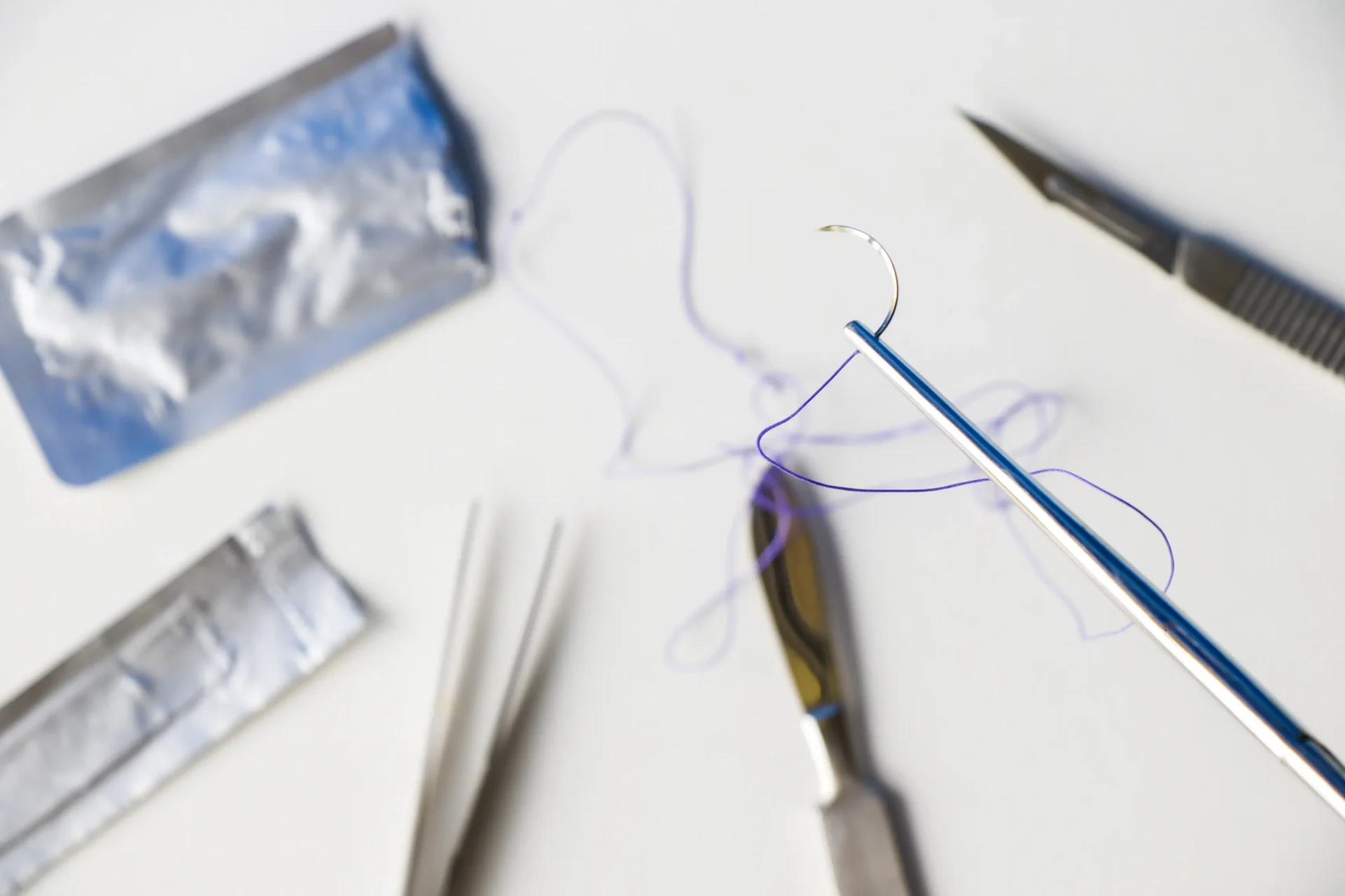 Close-up of surgical needle holder with curved suture needle and thread, surrounded by sterile medical instruments on white background