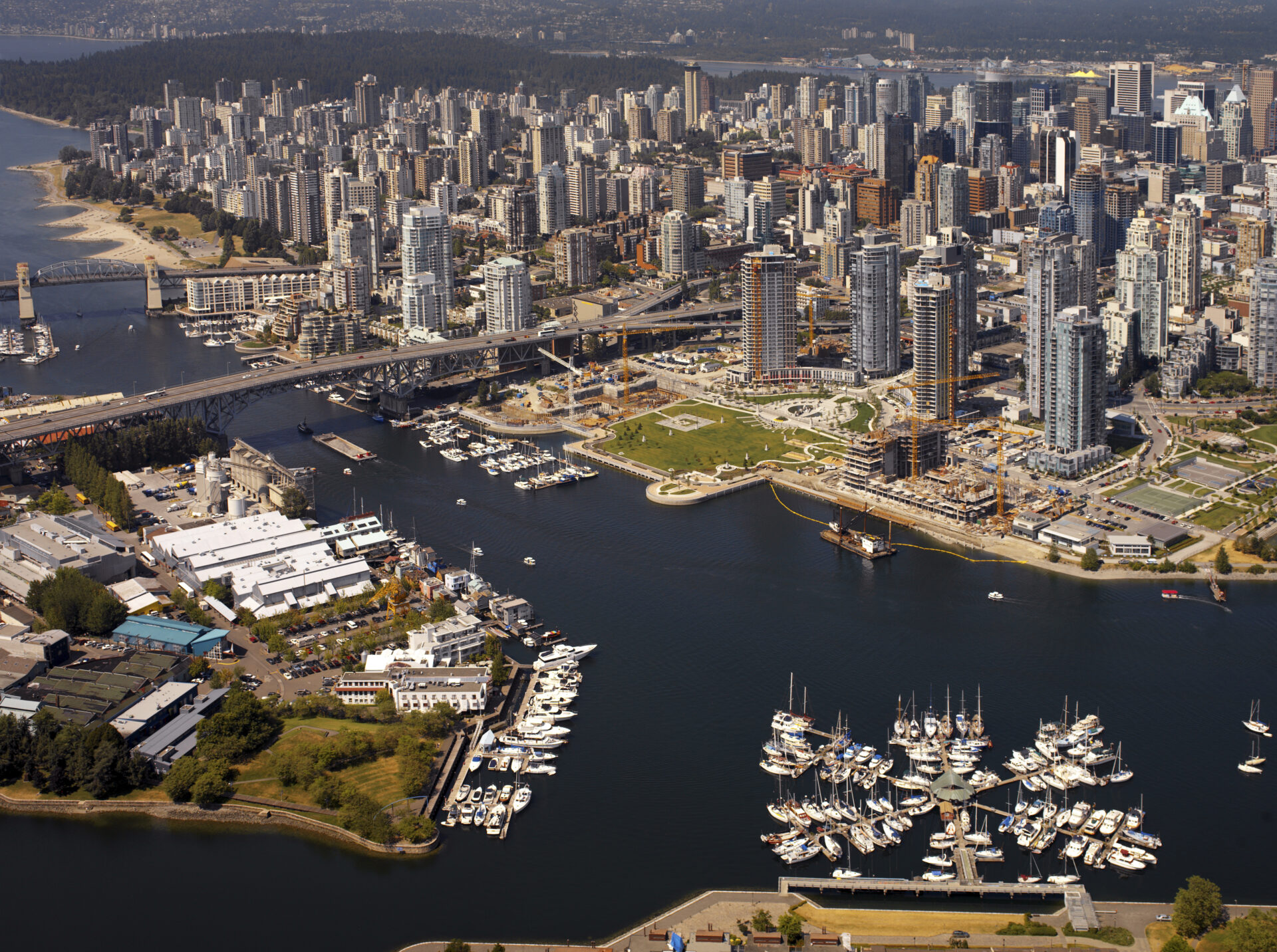 An aerial shot  of Vancouver BC captures a sprawling cityscape with numerous tall buildings and a harbor filled with boats. Multiple bridges span the waterway, connecting the urban center with lush, green landmasses.
